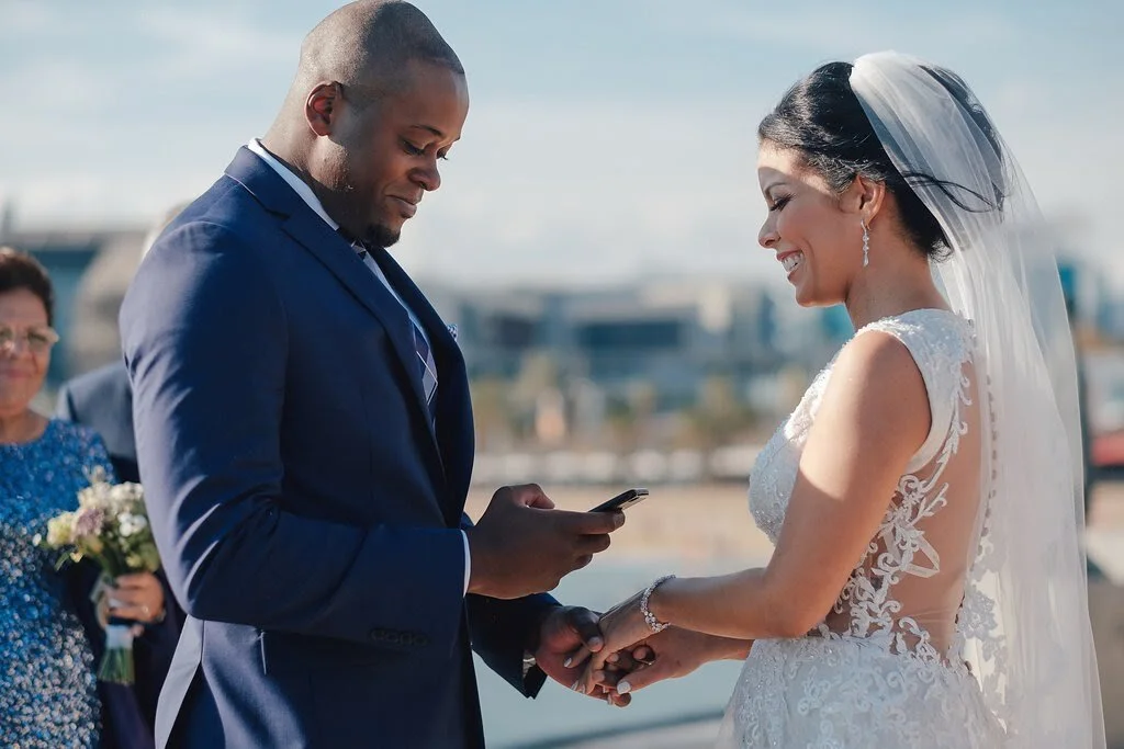 A bride and groom holding hands during their wedding ceremony outdoors, with the groom reading vows from a phone and the bride smiling in her wedding gown and veil.