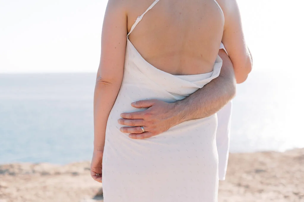 A couple standing on a beach, with the man's arm around the woman's waist and her arm hanging by her side, facing the ocean.