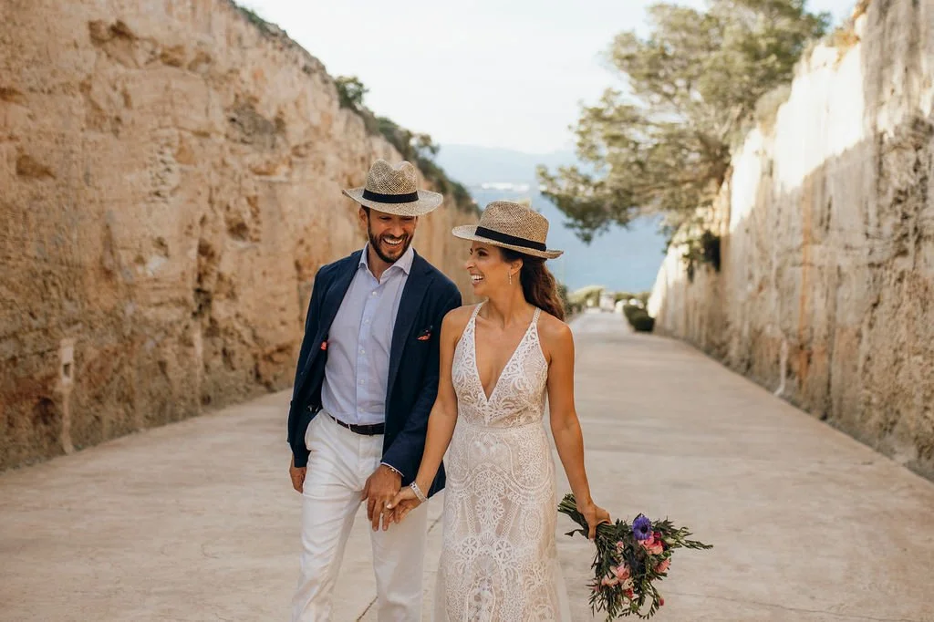 A couple in wedding attire walking outdoors on a sunny day, with rocky walls on either side and trees in the background, the woman holding a bouquet of flowers.