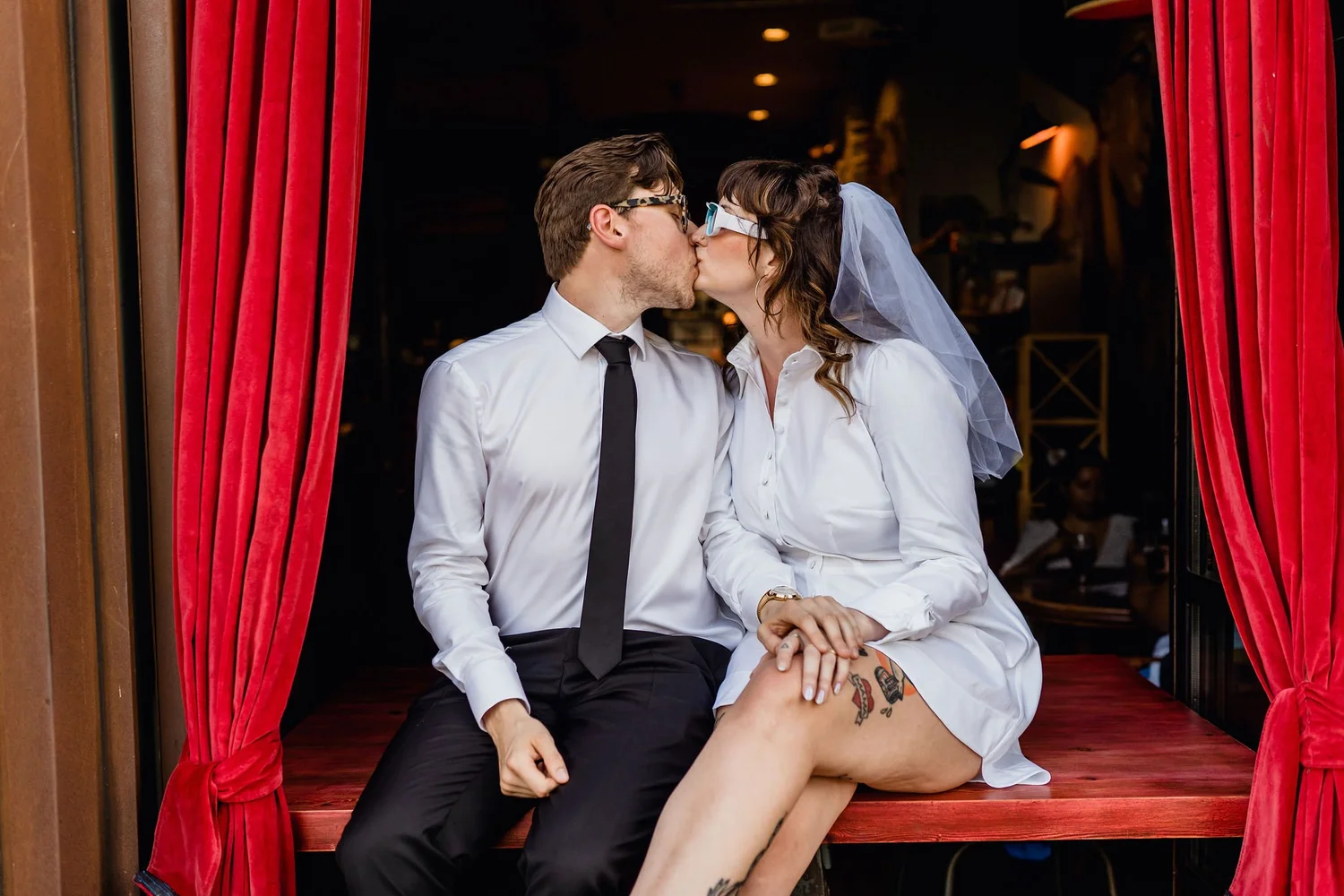 A man and woman dressed in wedding attire sharing a kiss, sitting in a red velvet theater box with red curtains, during a wedding celebration.