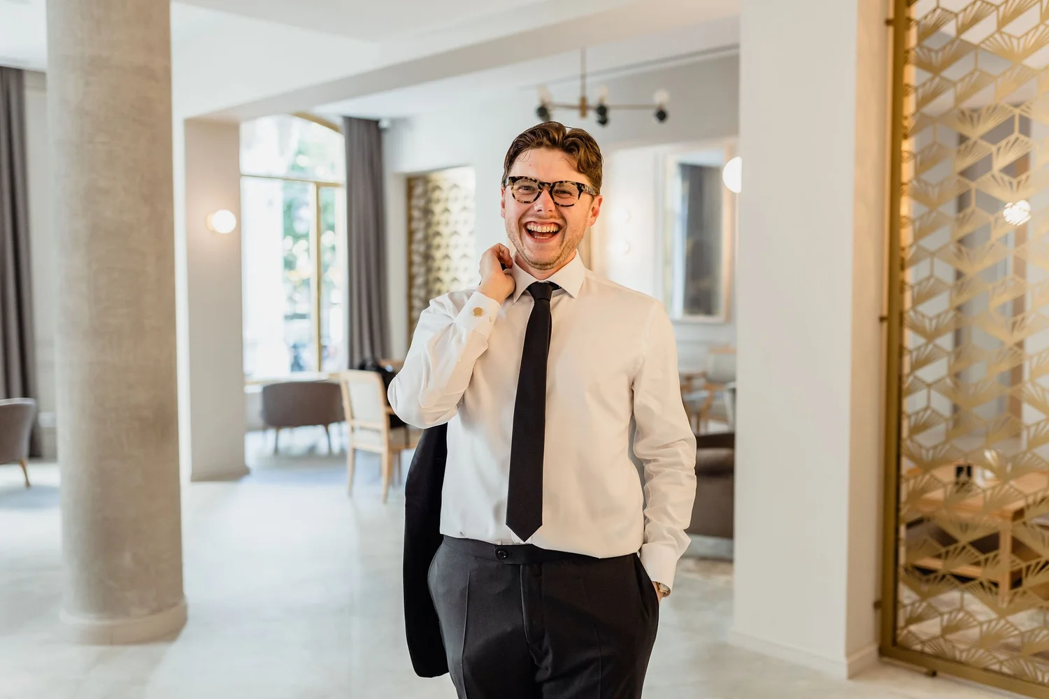 A young man in formal attire, wearing glasses and a black tie, is smiling and standing in a modern, well-lit interior space, possibly a restaurant or hotel lobby.