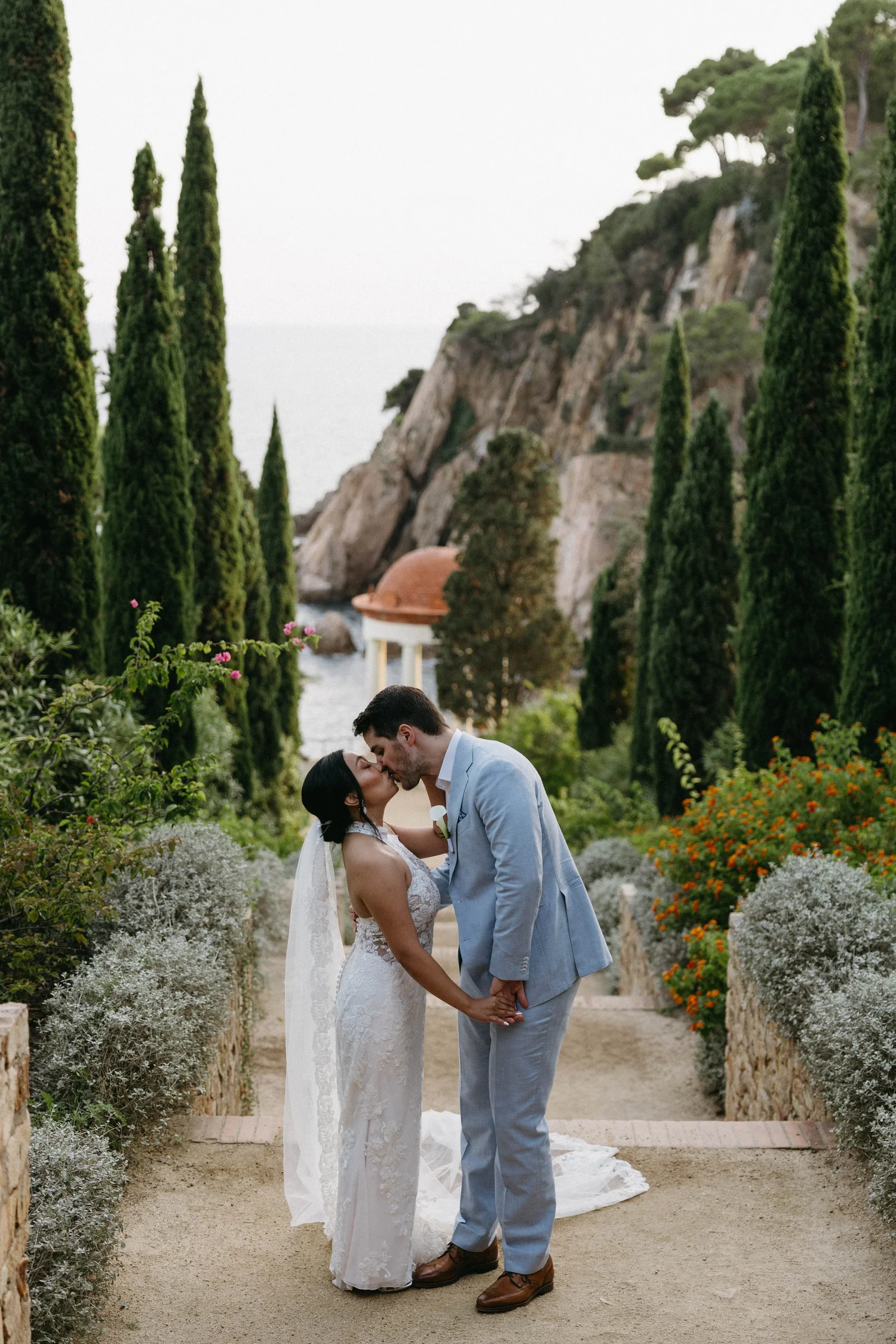 A bride and groom are kissing outdoors on a pathway bordered by lush green plants and colorful flowers, with a scenic hillside and water in the background during their wedding celebration.