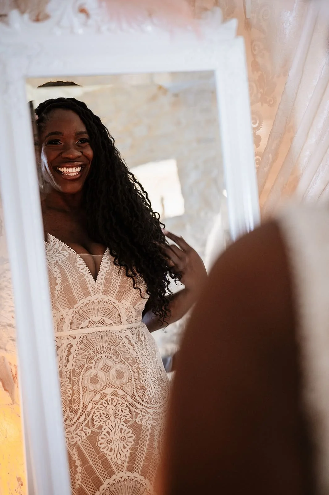 A woman with long, curly black hair smiling at herself in a mirror, wearing a cream-colored lace dress, in a room with warm lighting and decorative wallpaper.