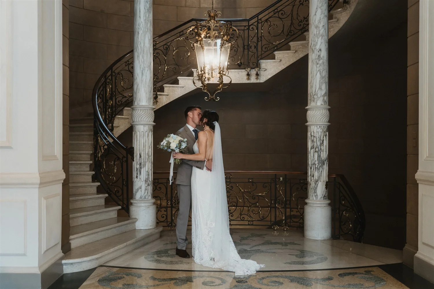 A bride and groom kissing inside a grand staircase hall with marble columns and an ornate chandelier overhead.