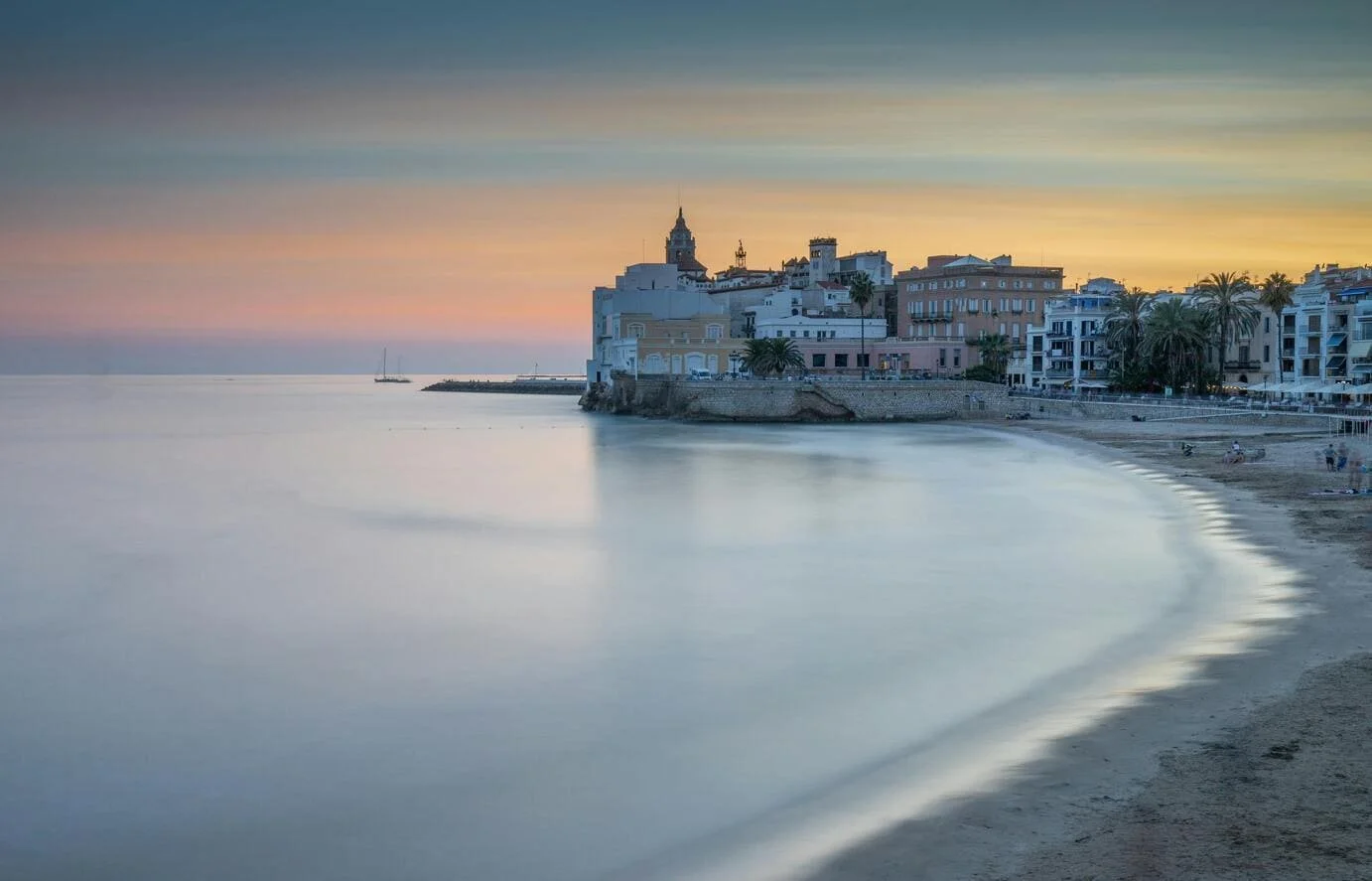 Beachside view of a historic town during sunset, with calm waters, sandy beach, colorful buildings, palm trees, and a church tower in the background.