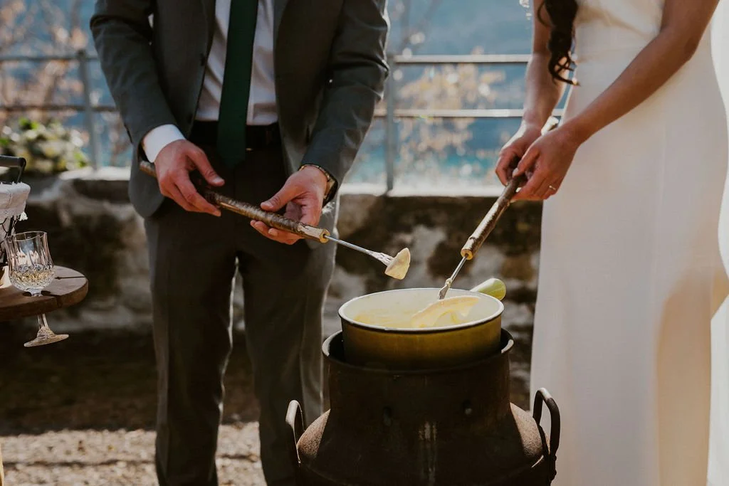 People reveal cheese fondue ceremony outdoors with a pot on a stand, using long forks to dip bread or other items into melted cheese.