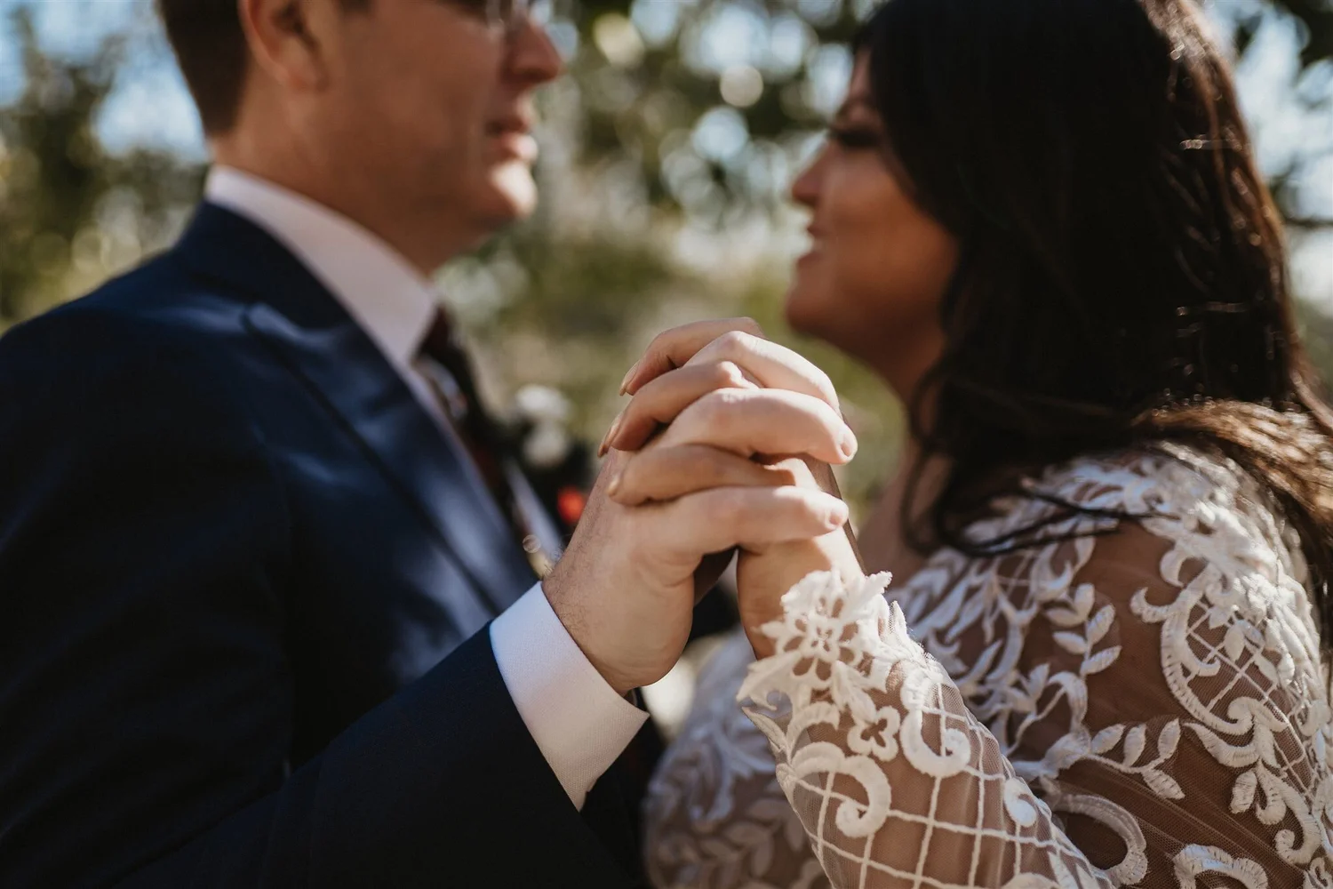 A man and woman holding hands close, facing each other outdoors with blurred trees in the background.