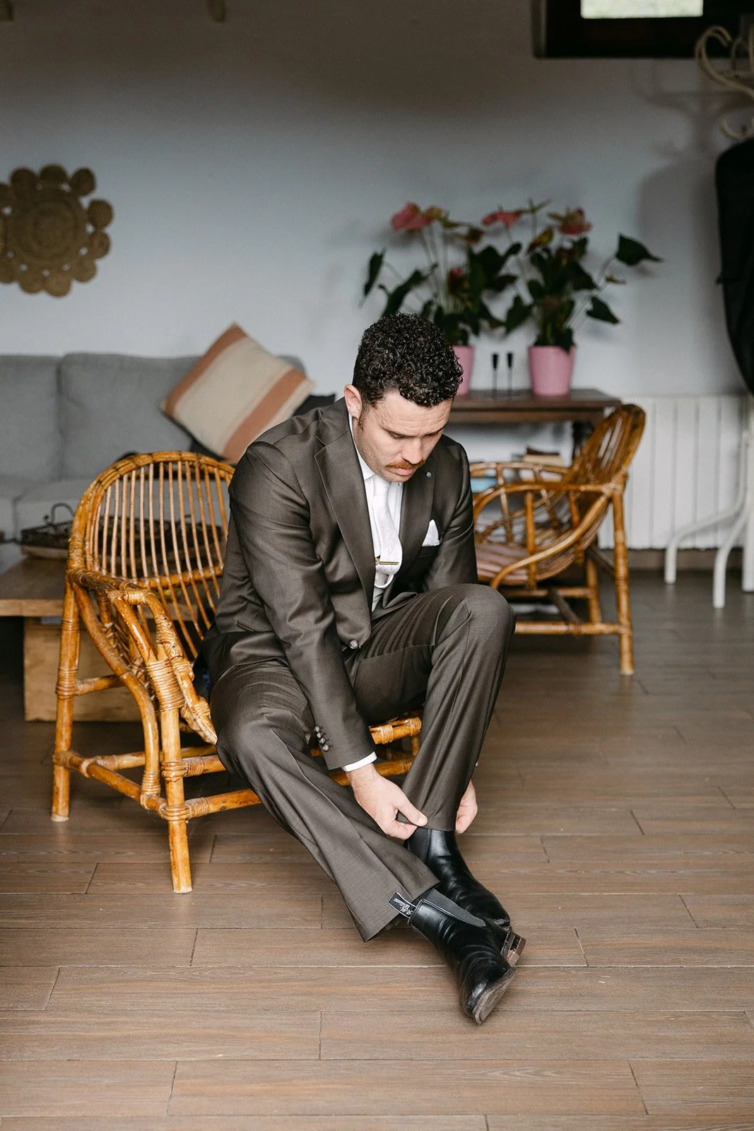 A man in a dark suit is sitting on a wicker chair, leaning forward, as he adjusts his black dress shoes. The room has wooden floors, a sofa with pillows, and a wooden table with pink potted plants in the background.