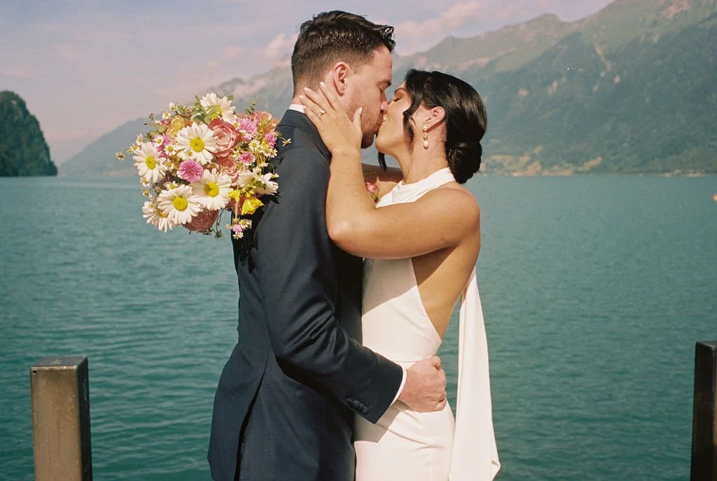 A bride and groom kiss by a lake with mountains in the background, the bride holding a bouquet of pink and white daisies.