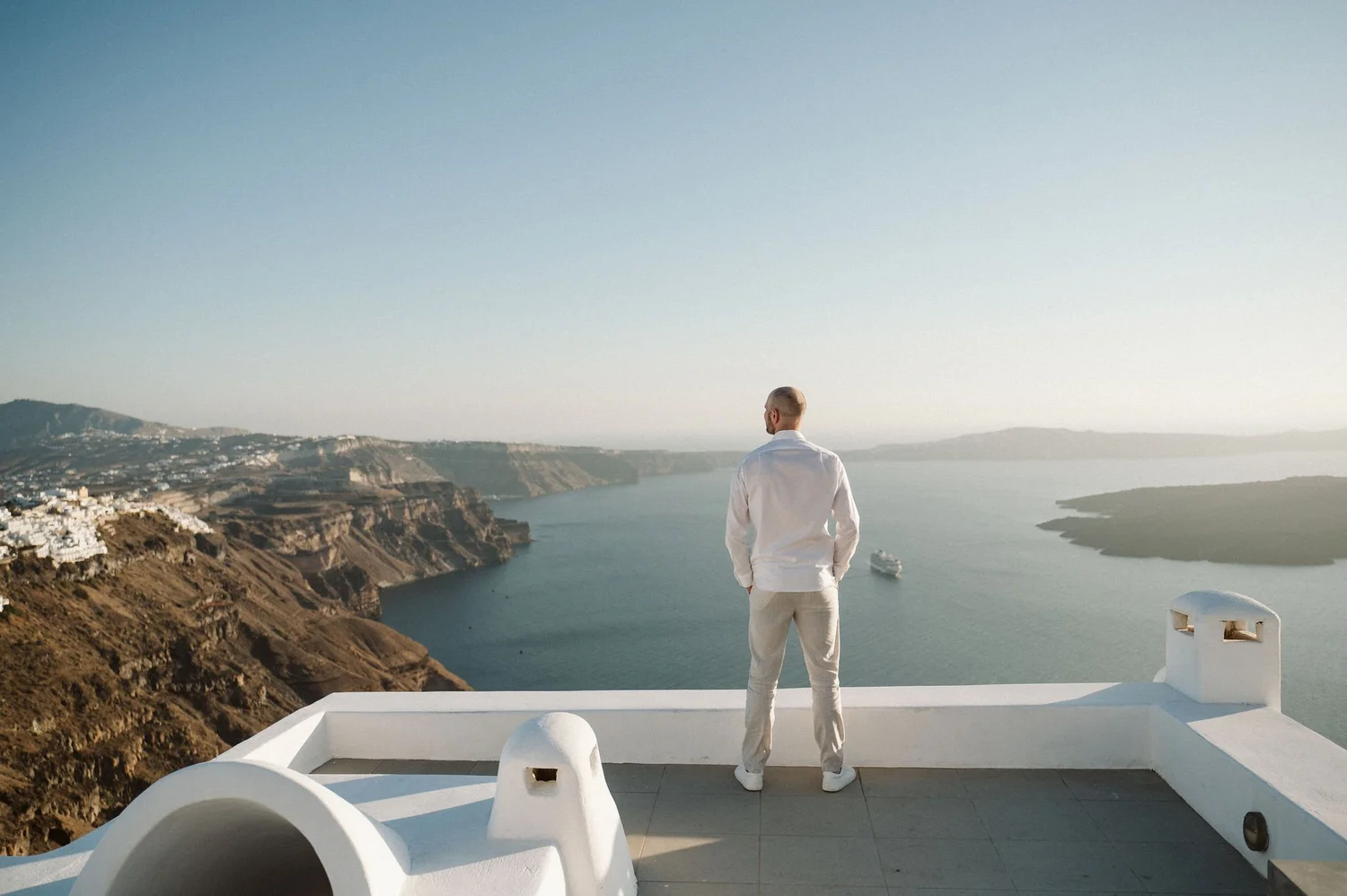A man in white clothing standing on a balcony overlooking the sea and coastal cliffs with white buildings, likely in a Mediterranean location.