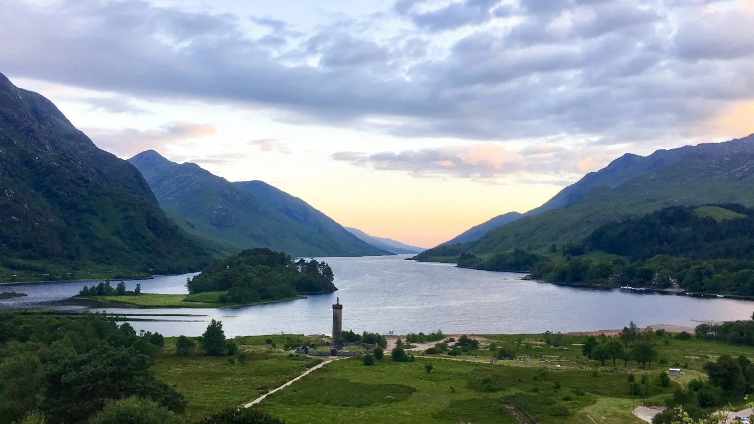 View of a river running through a lush green valley with mountains on both sides and a partly cloudy sky during sunset.