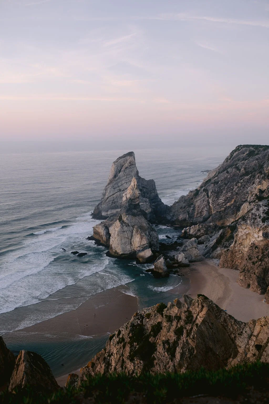 Scenic view of a rocky coastline at sunset with large sea stacks and waves crashing on the sandy beach below.