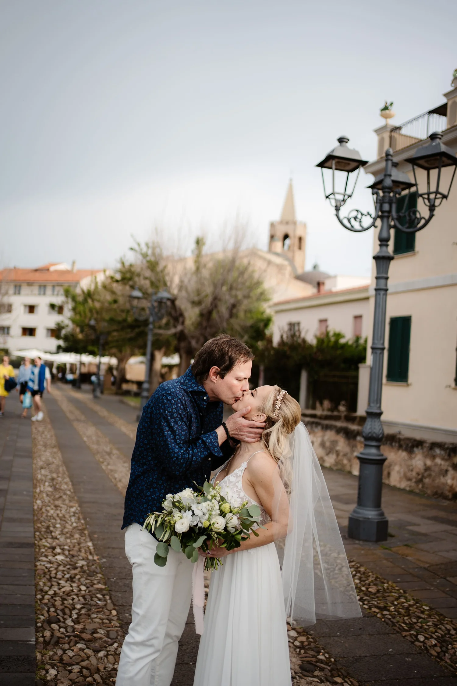 A couple on their wedding day sharing a kiss outdoors, with the man holding the woman by her face and her holding a bouquet of white flowers and greenery. They are standing on a pathway with a stone and gravel surface, and buildings, trees, a streetl