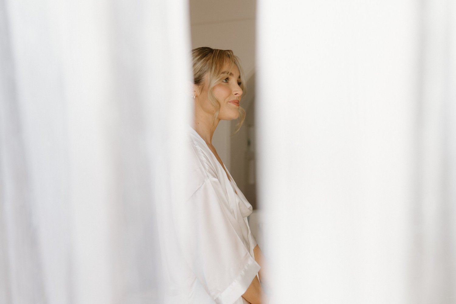 A woman with blonde hair styled in loose waves, wearing a white top, is seen smiling and looking to her right. The background shows a neutral interior with soft lighting.