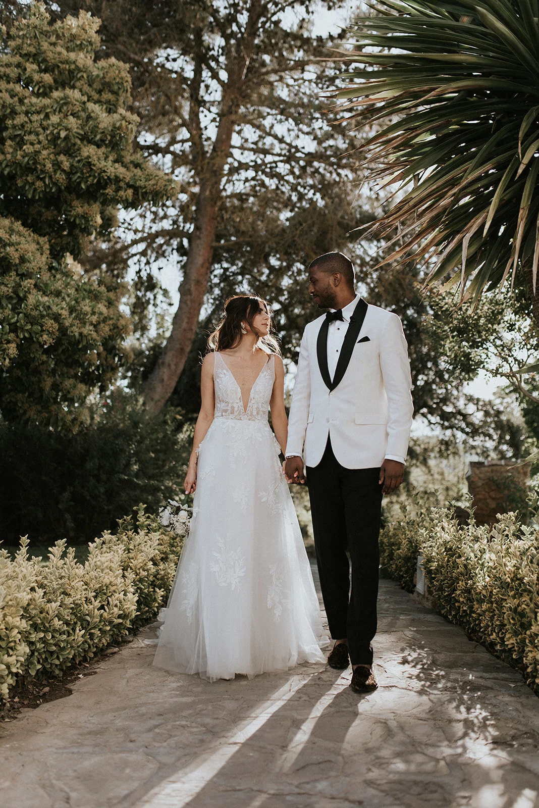 A bride and groom holding hands outdoors, walking on a stone pathway surrounded by greenery and trees, during a wedding photoshoot.