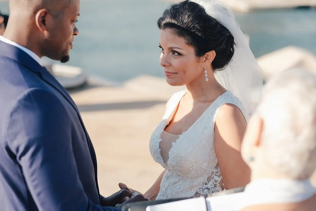 Bride and groom at their wedding ceremony by the water, holding hands and gazing at each other.