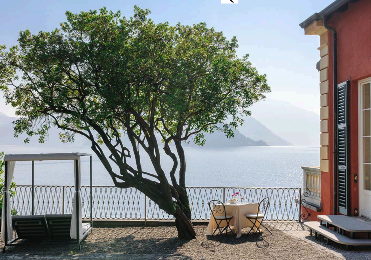 A lakeside outdoor patio with a large tree, a swing, a table with two chairs by the railing overlooking the water, and part of a building with a red exterior and black shutters.