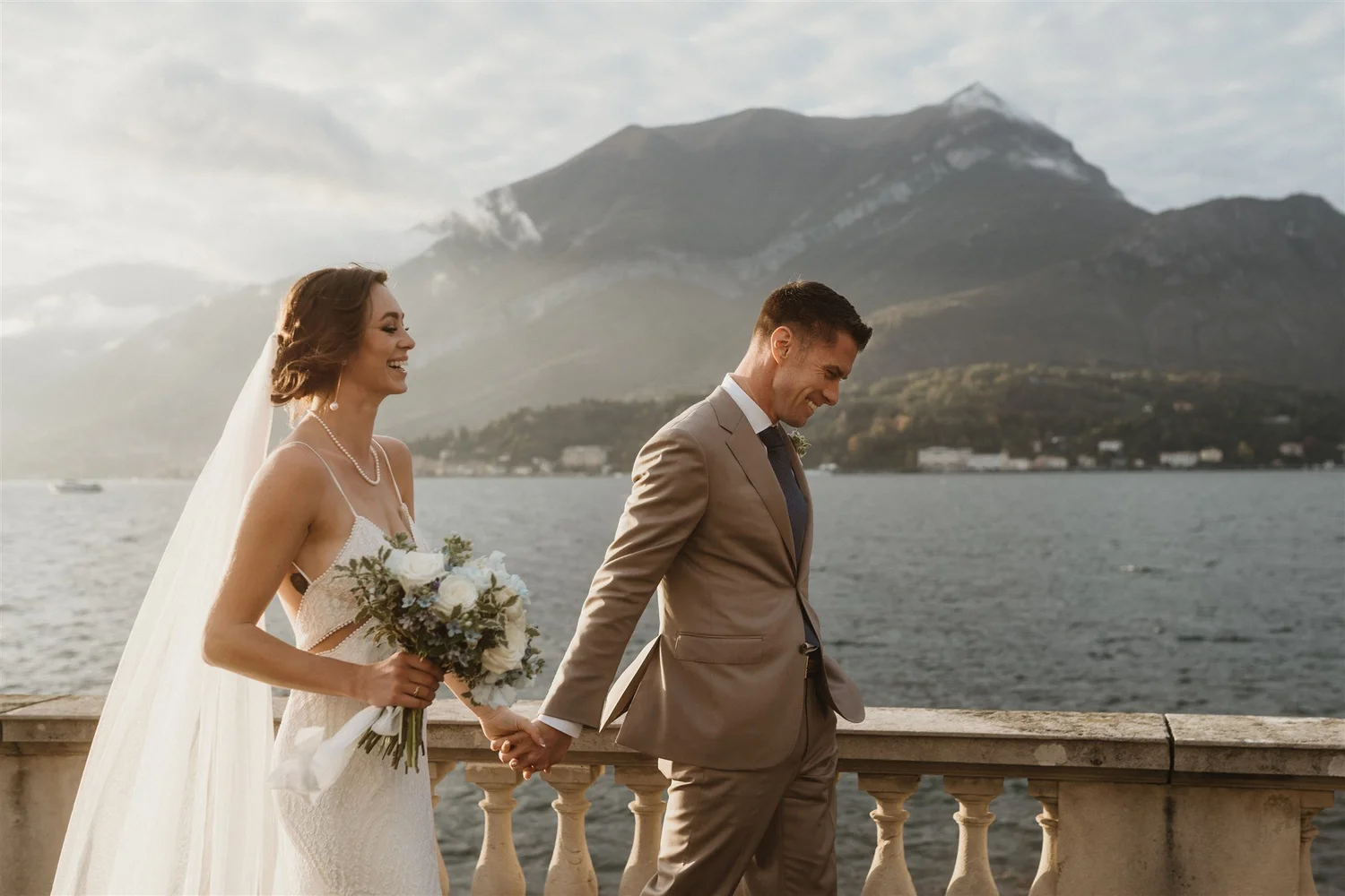 A bride and groom holding hands and walking along a waterfront with a mountain in the background, during their wedding celebration.