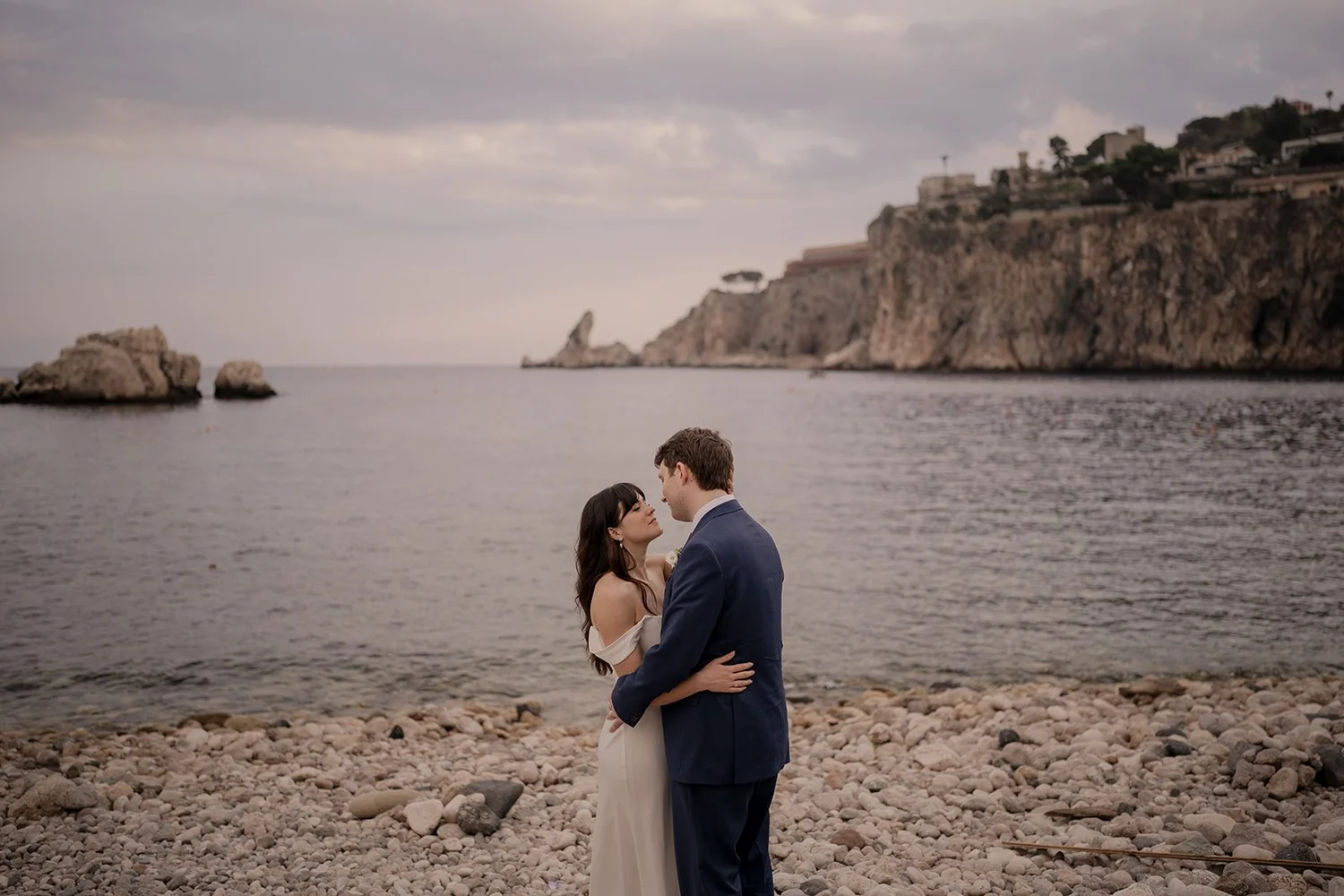 A couple in wedding attire stands on a rocky beach, embracing and gazing into each other's eyes, with a large rocky cliff and buildings in the background over the water.