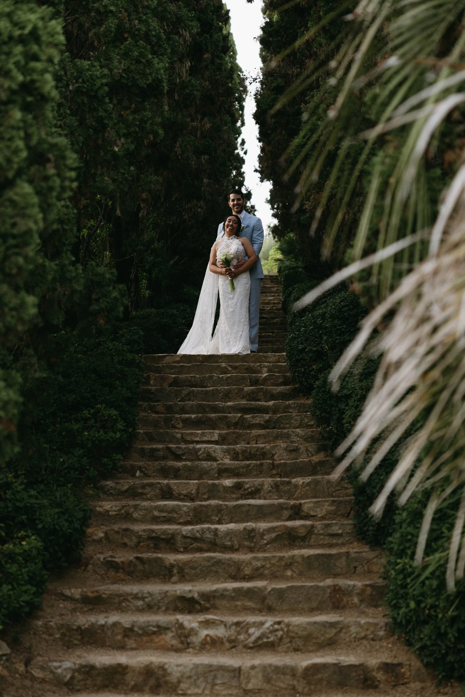 A newlywed couple is standing on a stone staircase surrounded by dense green foliage, with the bride holding a bouquet of white flowers and smiling, while the groom stands behind her, dressed in a light blue suit, smiling.