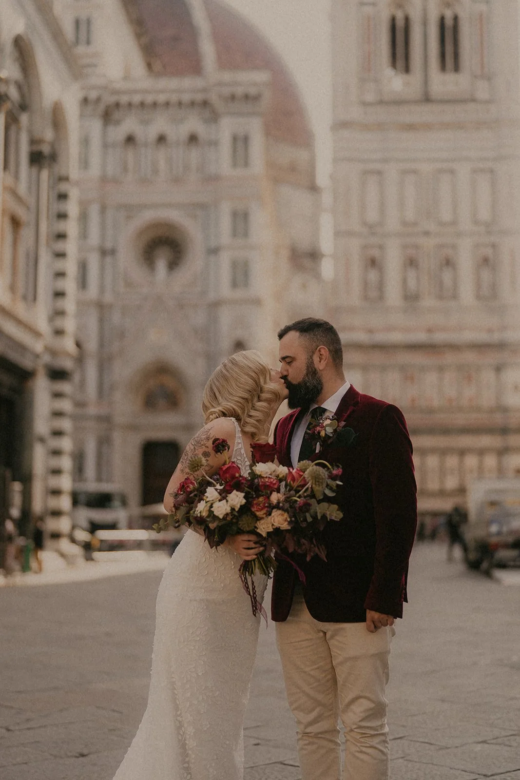 A newlywed couple sharing a kiss, holding a large bouquet of flowers in front of a historic European building.
