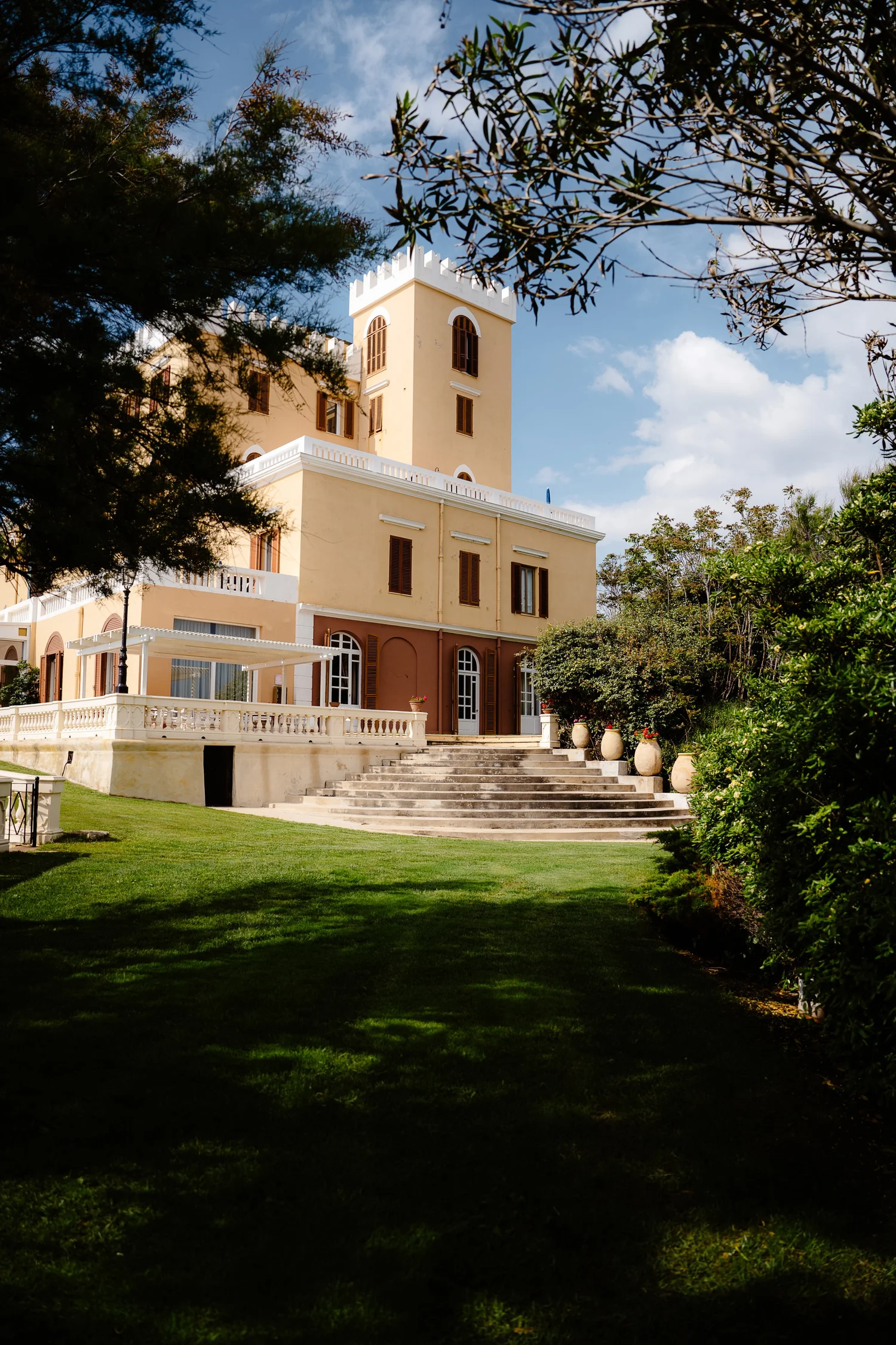 A yellow castle-like building with a tower, surrounded by trees and a well-maintained lawn, under a partly cloudy sky.