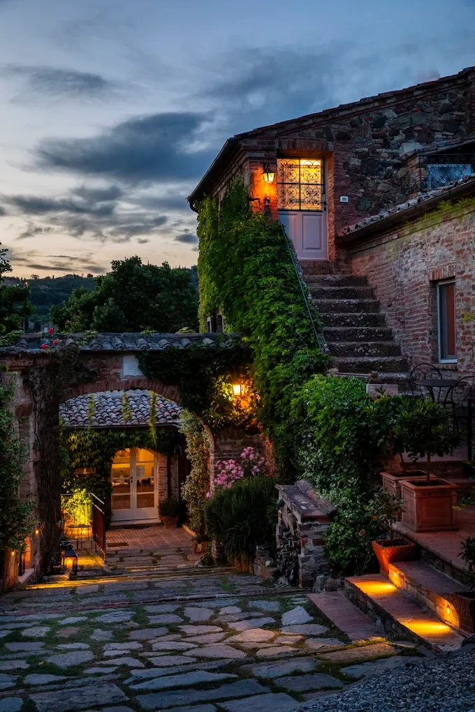 A rustic stone house with ivy-covered walls, outdoor lighting, and a stone pathway leading to a door, set against a cloudy evening sky.
