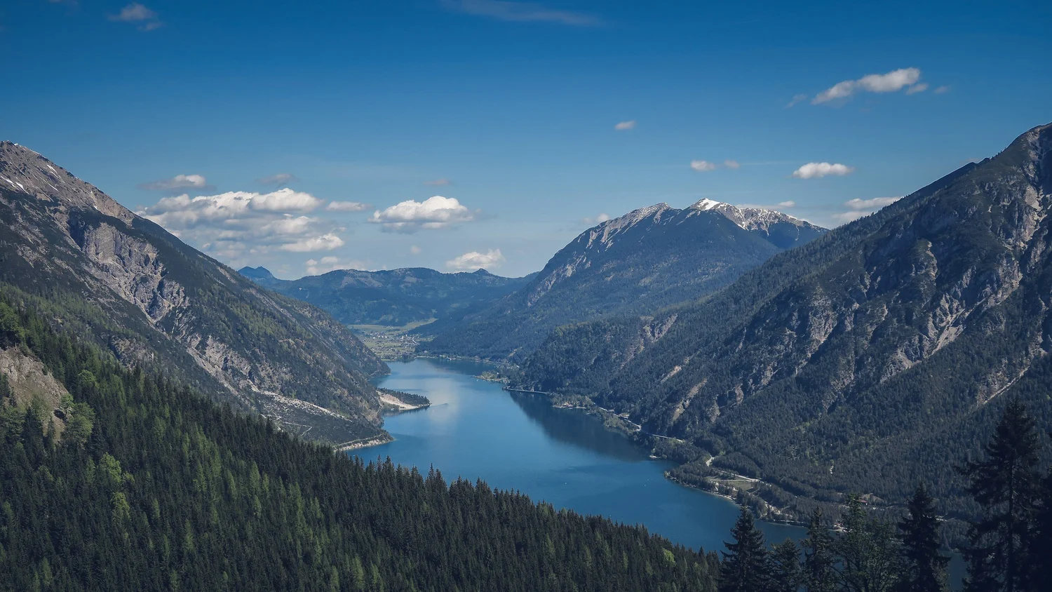 A scenic view of a mountain valley with a river winding between forested mountains and snow-capped peaks under a partly cloudy blue sky.