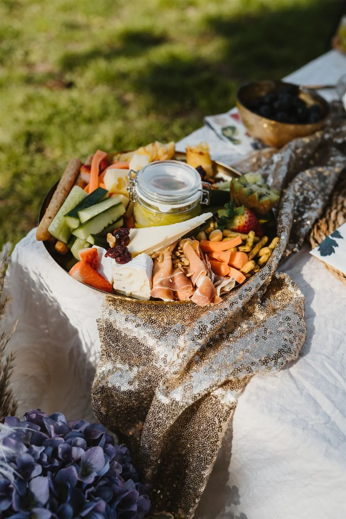 A charcuterie board filled with assorted cheeses, sliced cucumbers, strawberries, grapes, prosciutto, nuts, and a jar of mustard, set on a decorated table outdoors with a grassy background.