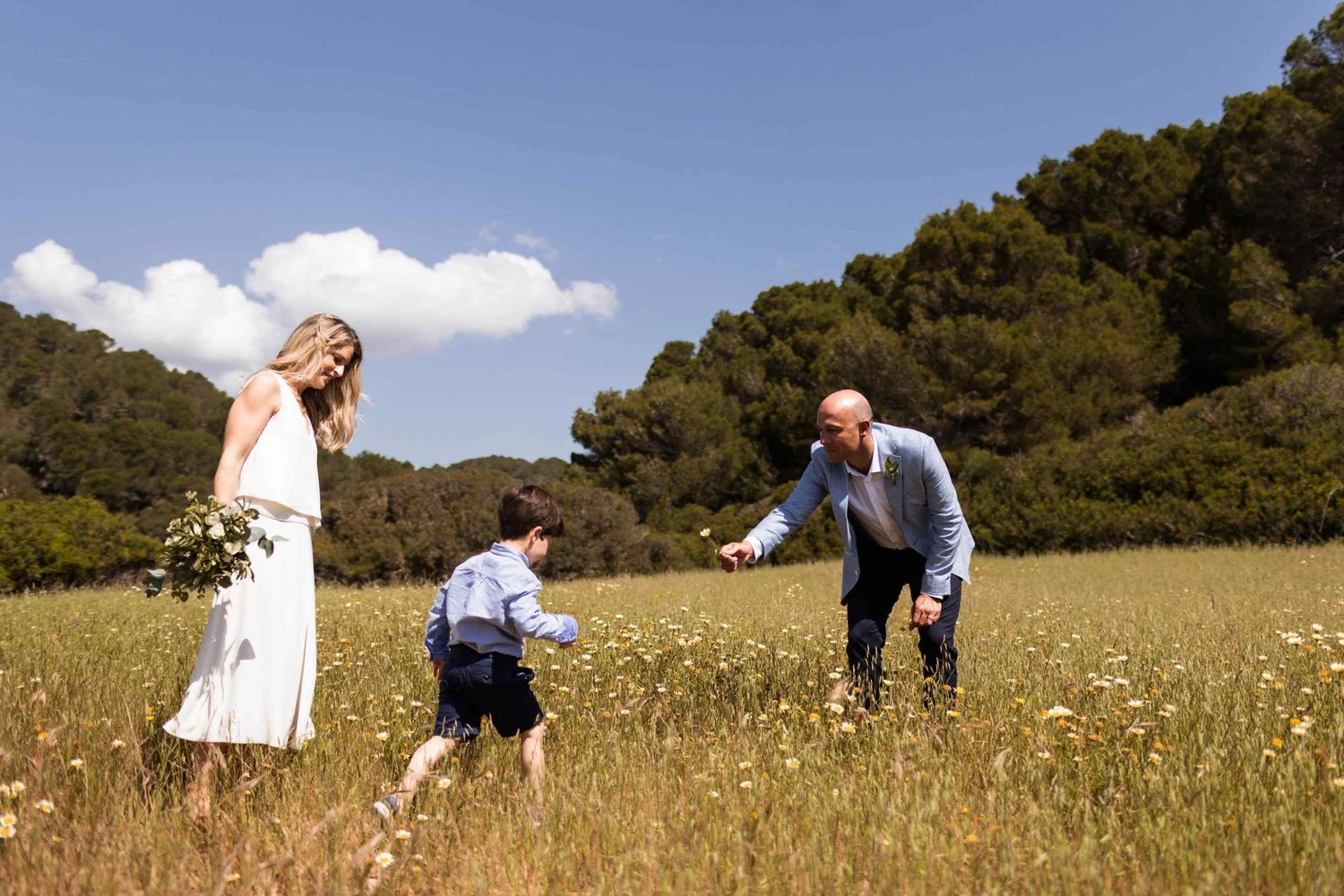 A family having a picnic in a grassy field surrounded by trees under a blue sky with white clouds. The woman is in a white dress holding a bouquet, and the man and two boys are picking flowers, dressed in light blue shirts.