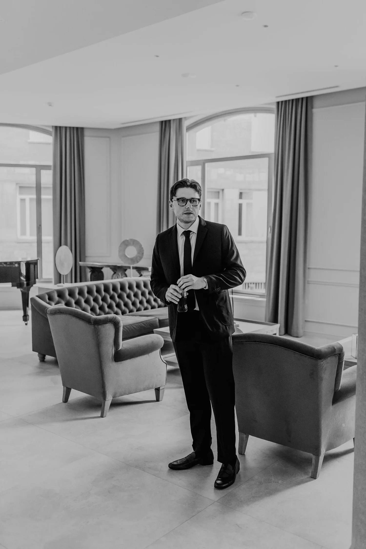 A man in a suit and tie stands in a modern living room holding a glass. The room features large windows with curtains, a piano, and multiple chairs and sofas.