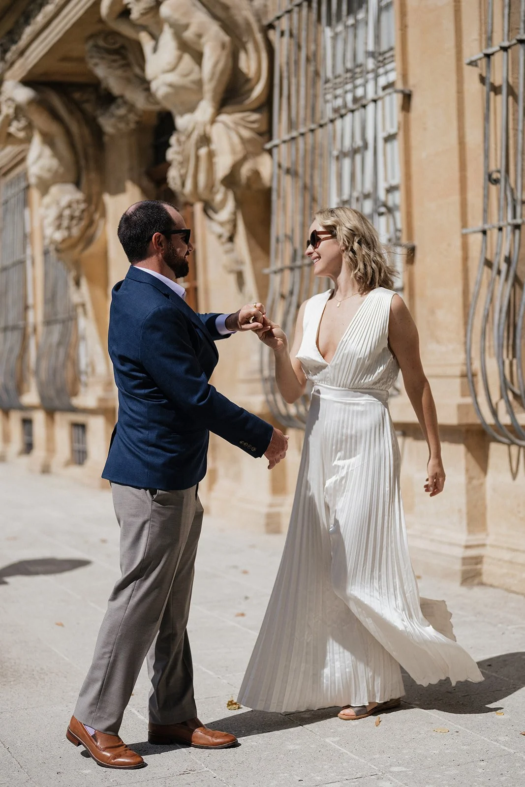 A couple is dancing and smiling outside in front of a historic building, with the man wearing a dark blazer and light trousers, and the woman wearing a white, pleated dress and sunglasses.