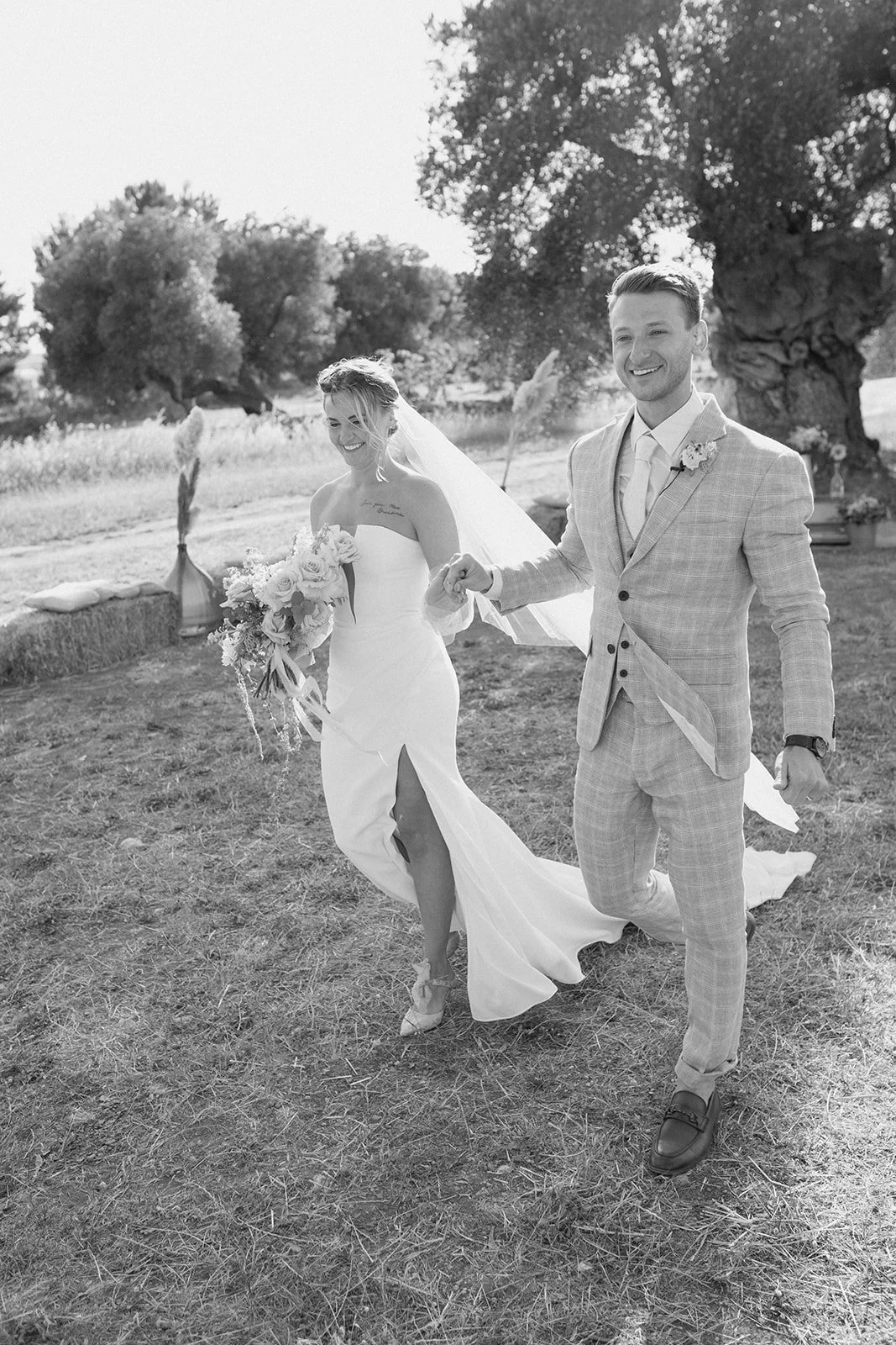 Black and white photo of a bride in a strapless wedding dress holding a bouquet, and a groom in a light-colored plaid suit, walking hand in hand outdoors on a sunny day with trees in the background.
