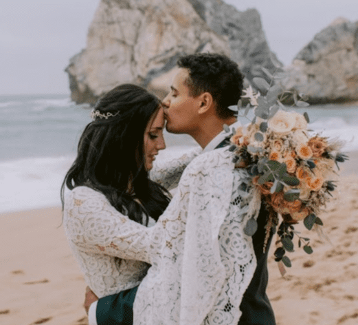 A couple dressed in wedding attire embrace on a beach, with the groom kissing the bride's forehead and holding a bouquet of flowers, rocky formations in the background.