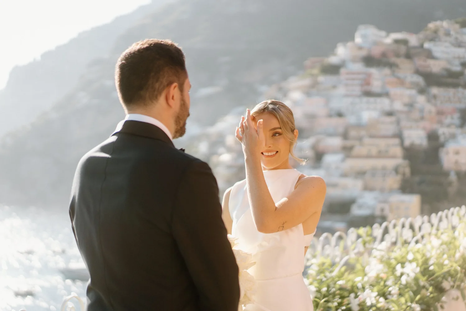 A bride and groom during their wedding ceremony outdoors, with the groom facing the bride and the bride smiling and touching her forehead with her hand, set against a scenic background of hills and houses.