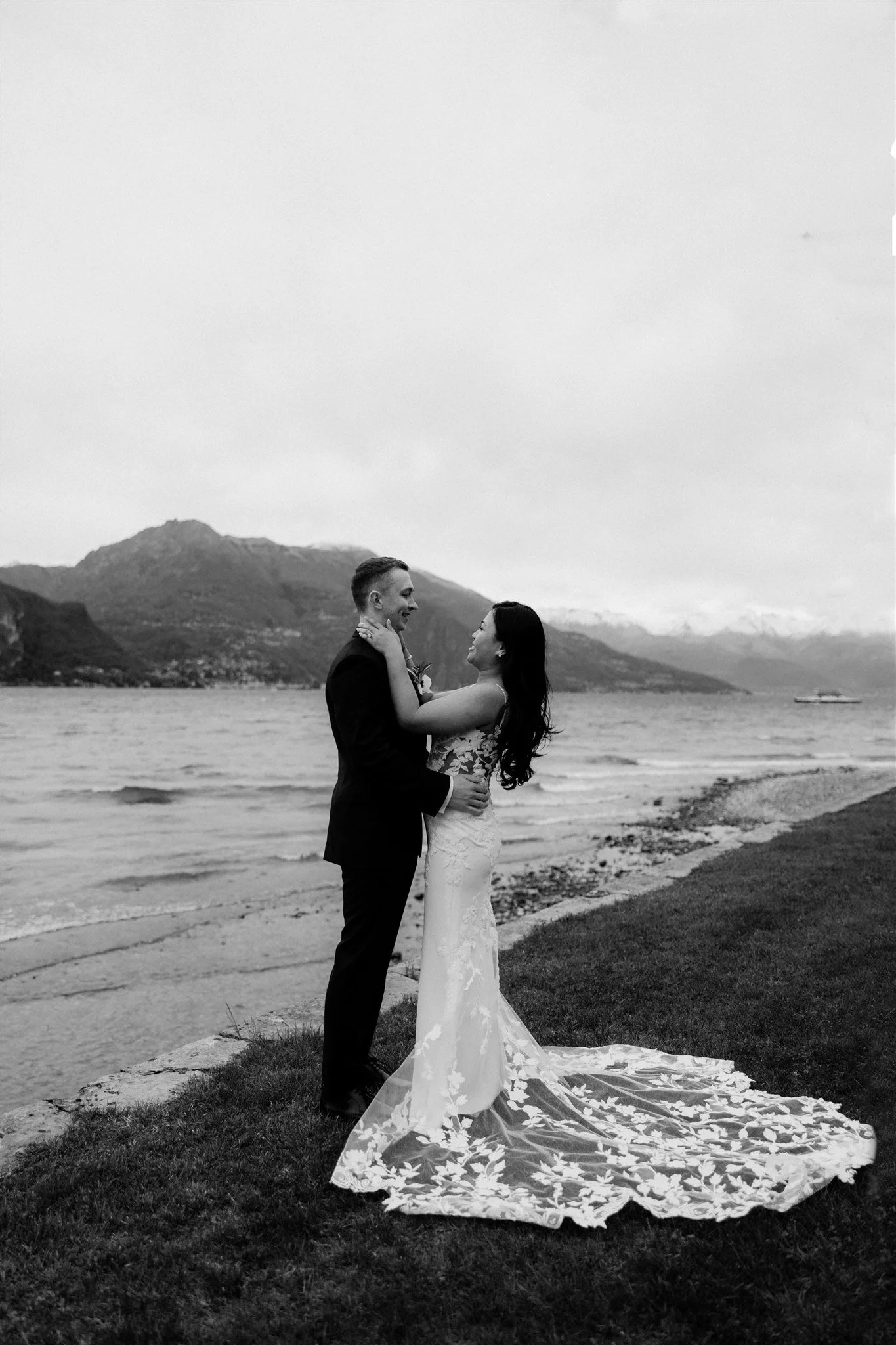 A black and white photo of a bride and groom holding each other and smiling on a beach with mountains in the background, the bride wearing a lacy wedding dress with a long train and the groom in a dark suit.