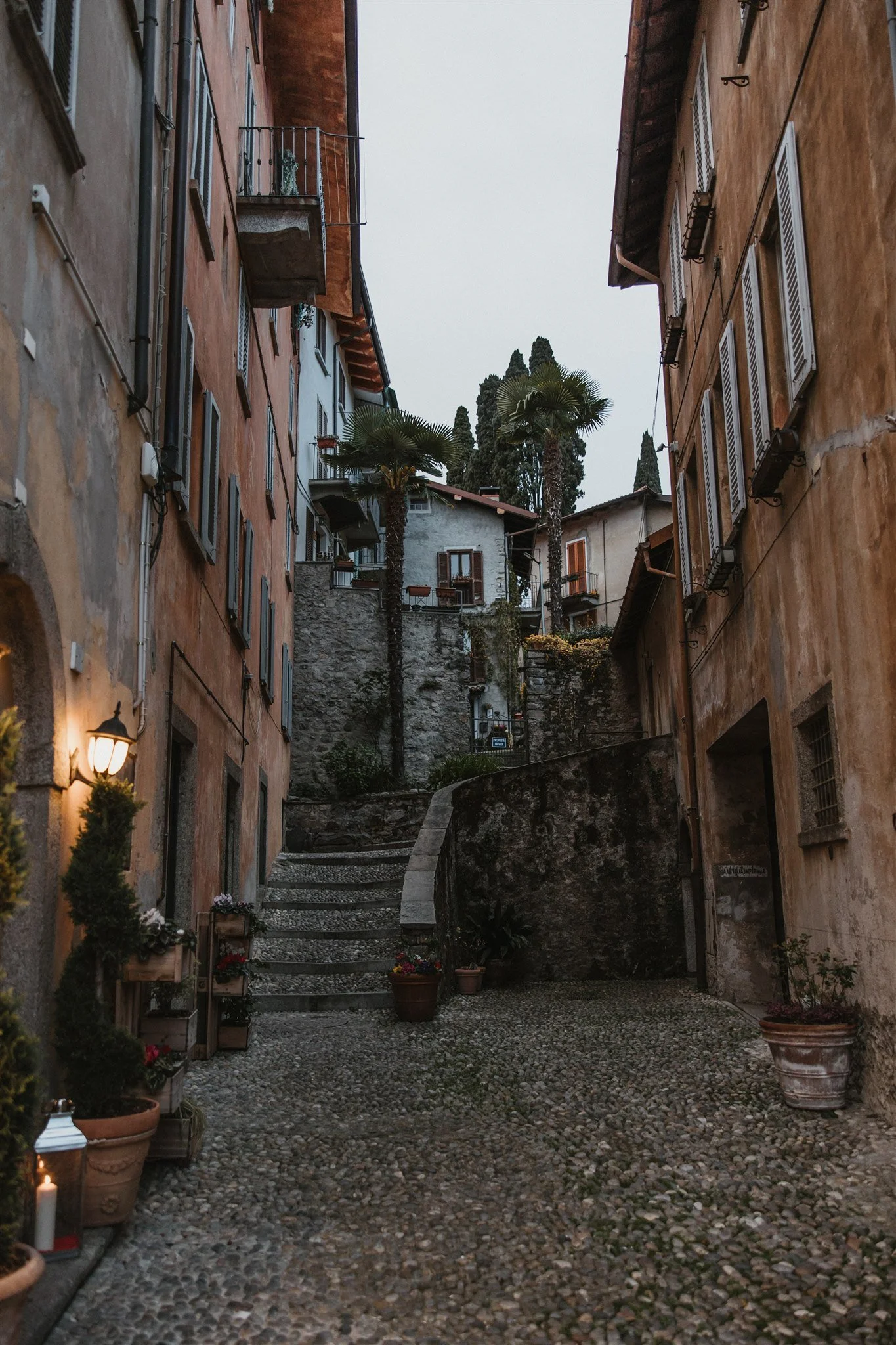 Cobblestone alleyway between tall, colorful buildings with flowers, potted plants, and lit candles along the sides, leading to stairs and trees in the distance.