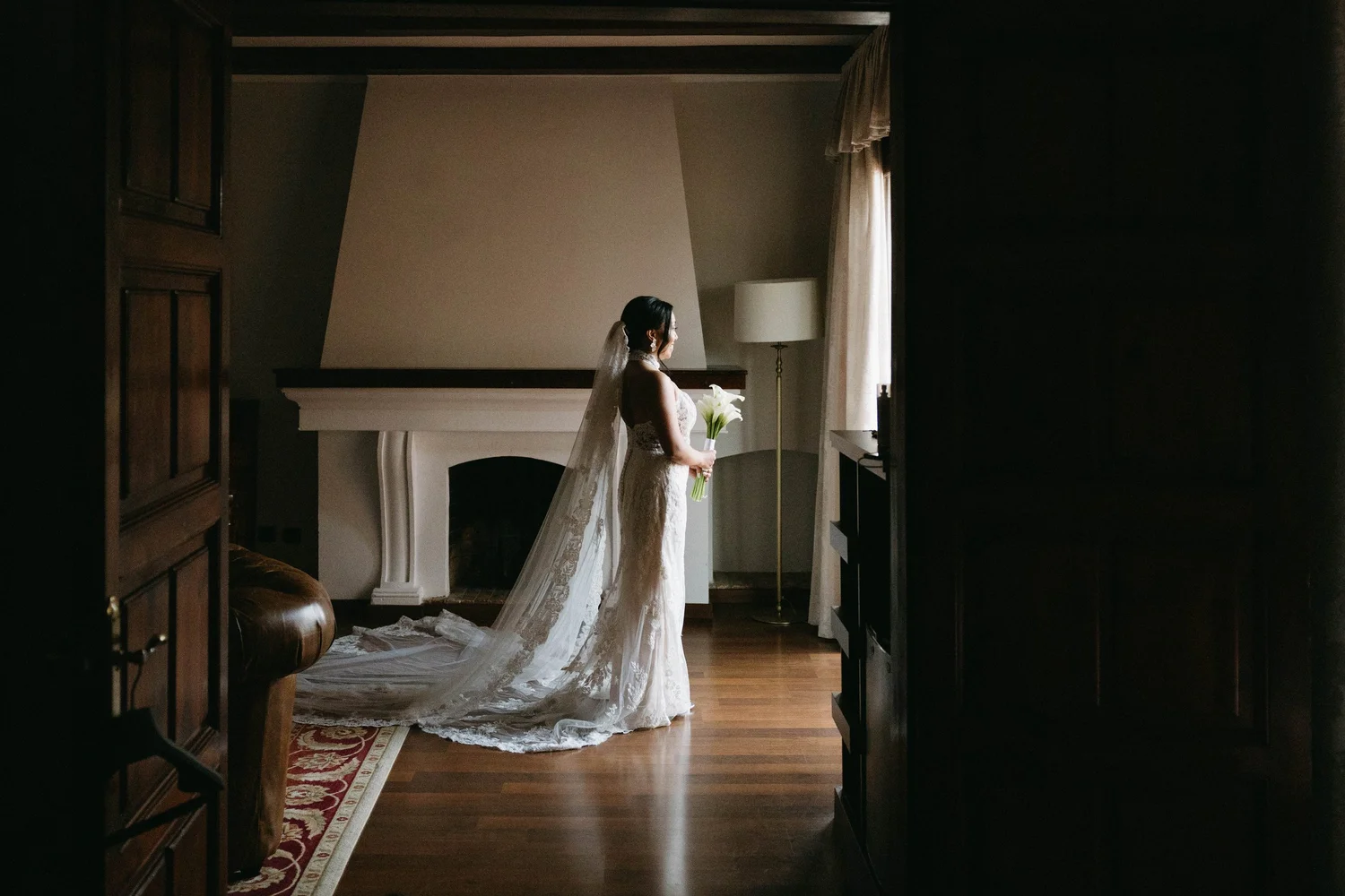 A bride stands in a dimly lit room holding a bouquet of white lilies, wearing a lace wedding dress with a long train and veil, looking out a window.