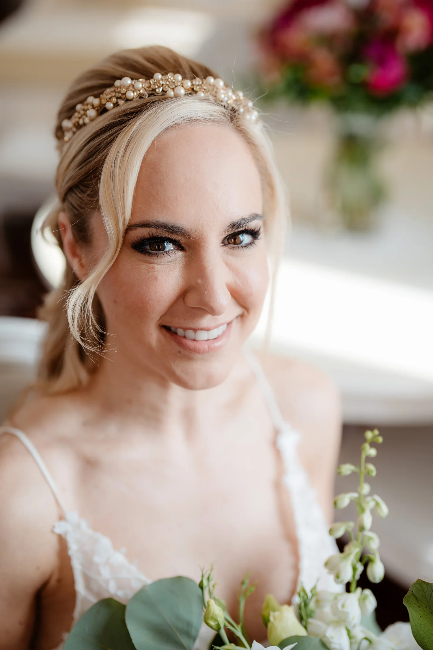 A smiling woman with blonde hair styled in loose waves, wearing a pearl-embellished headband and white lace spaghetti-strap dress, holding white flowers and greenery, with a blurred background featuring pink flowers.