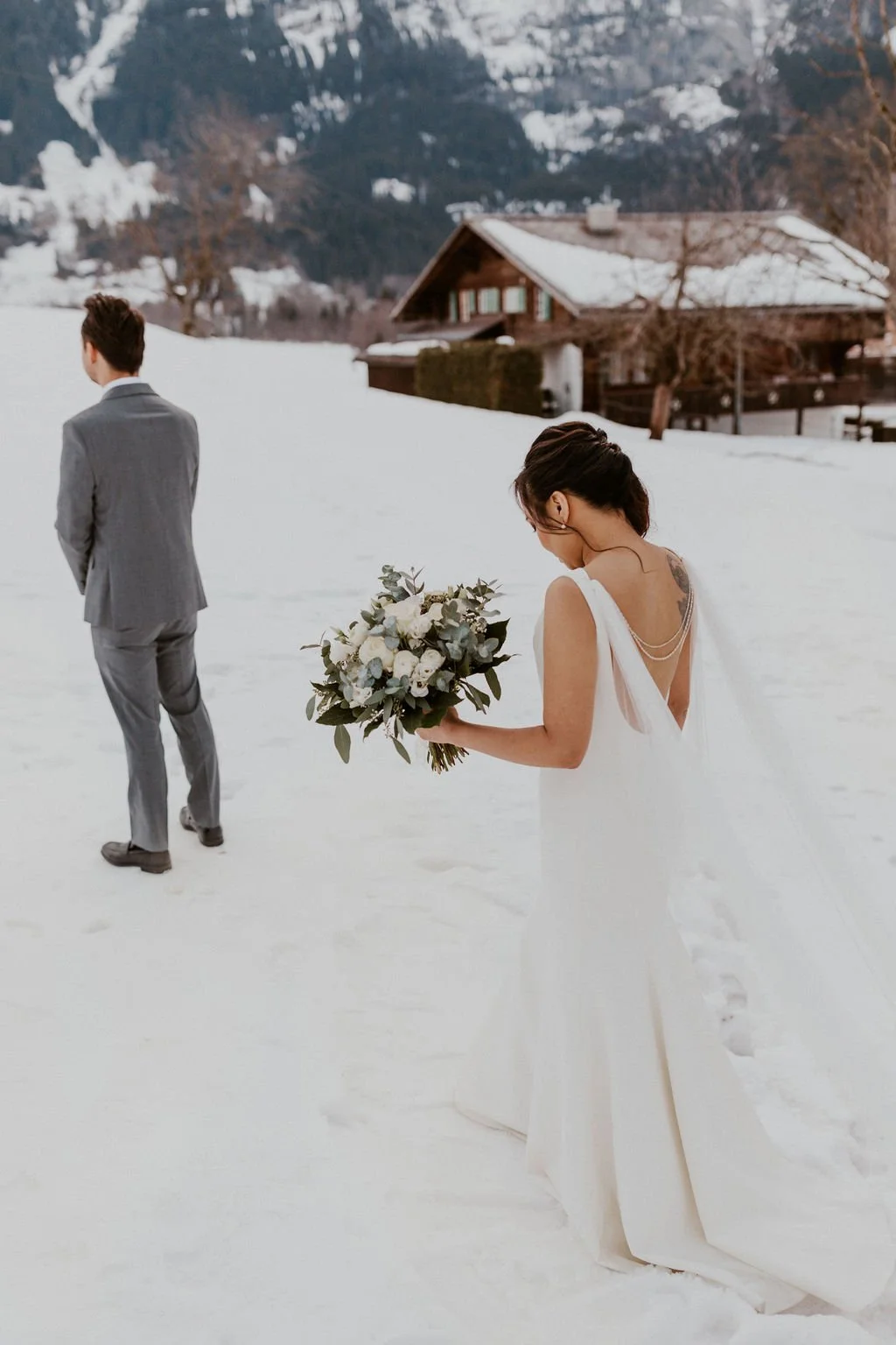A bride in a white wedding dress holding a bouquet looking down in the snow while a groom in a gray suit walks away in a snowy outdoor setting with a mountain and house in the background.