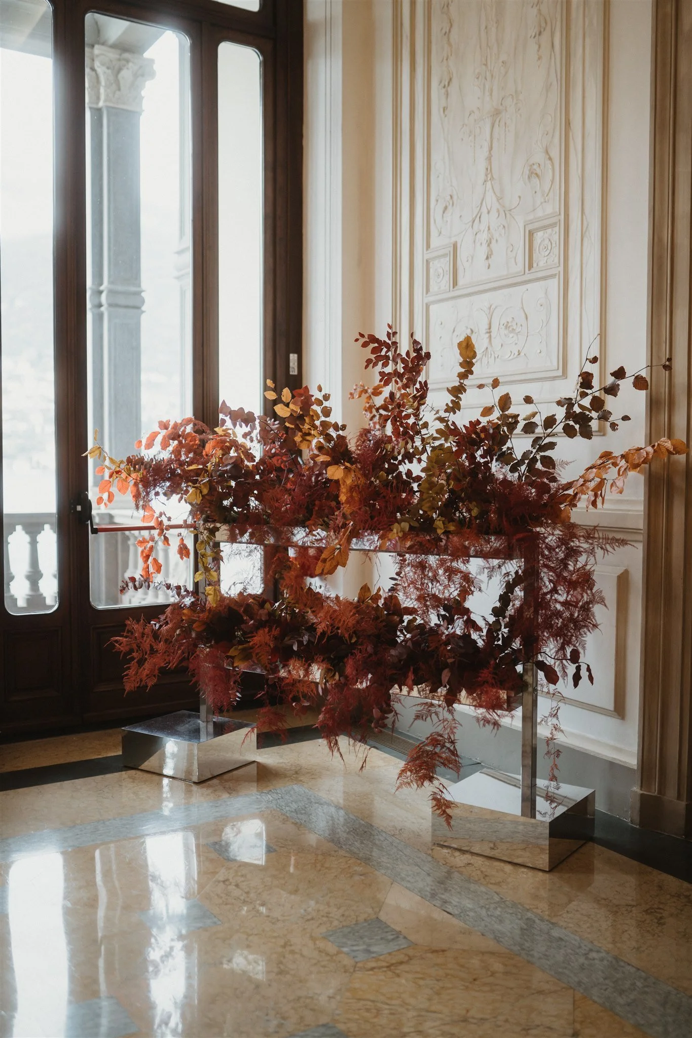 An ornamental floral arrangement featuring red and brown leaves in front of an elegant wall with intricate moldings, next to a large window with wooden frames.
