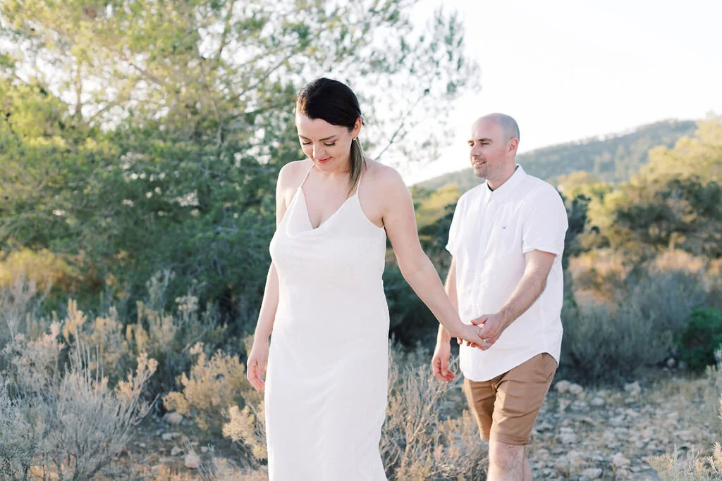 A woman in a white dress and a man in a white shirt and beige shorts holding hands in a natural outdoor setting with trees and hills.