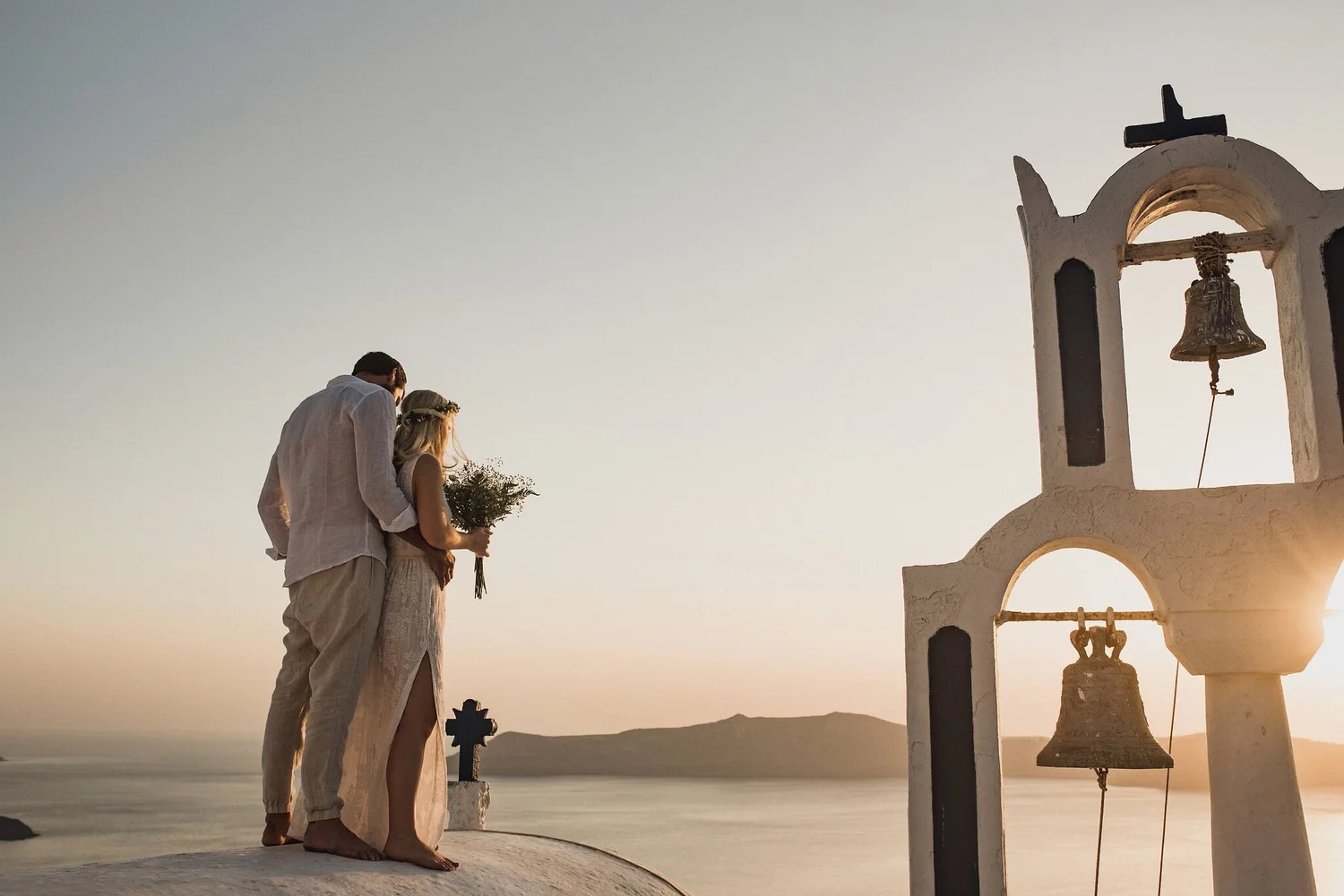 A couple standing on a white religious building rooftop during sunset, with mountains and water in the background, near a bell tower with a cross and bells.