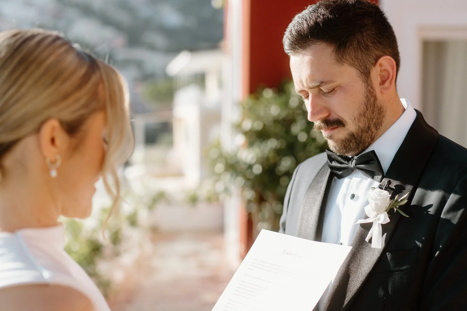 A groom in a black tuxedo and bow tie appears to be emotional during a wedding ceremony, possibly reading vows. The bride, with blonde hair, is visible from behind, standing opposite him.