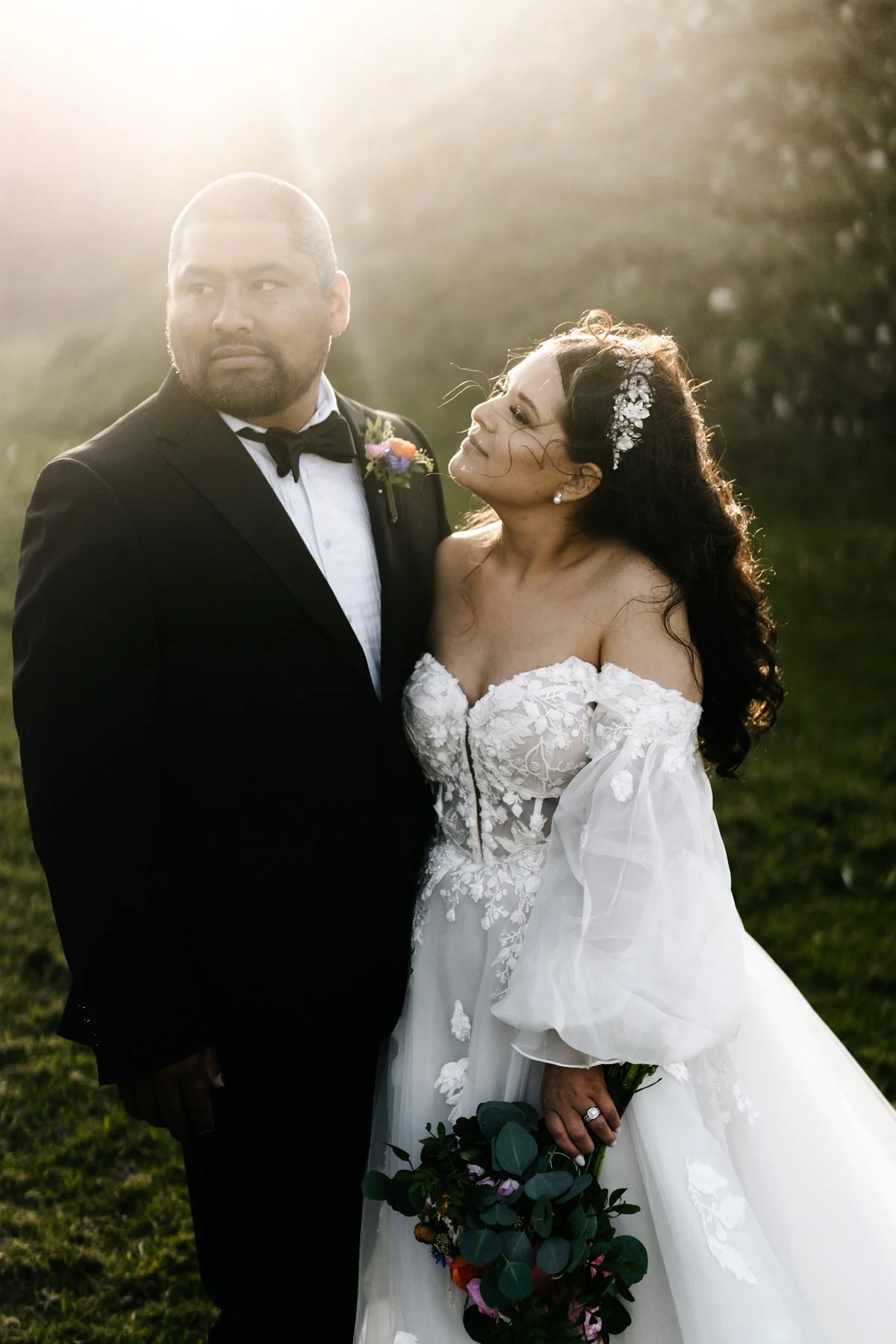 A bride and groom posing outdoors during sunset, the bride in a white wedding gown holding a bouquet, the groom in a black tuxedo with a bow tie, both standing on grass with trees in the background.