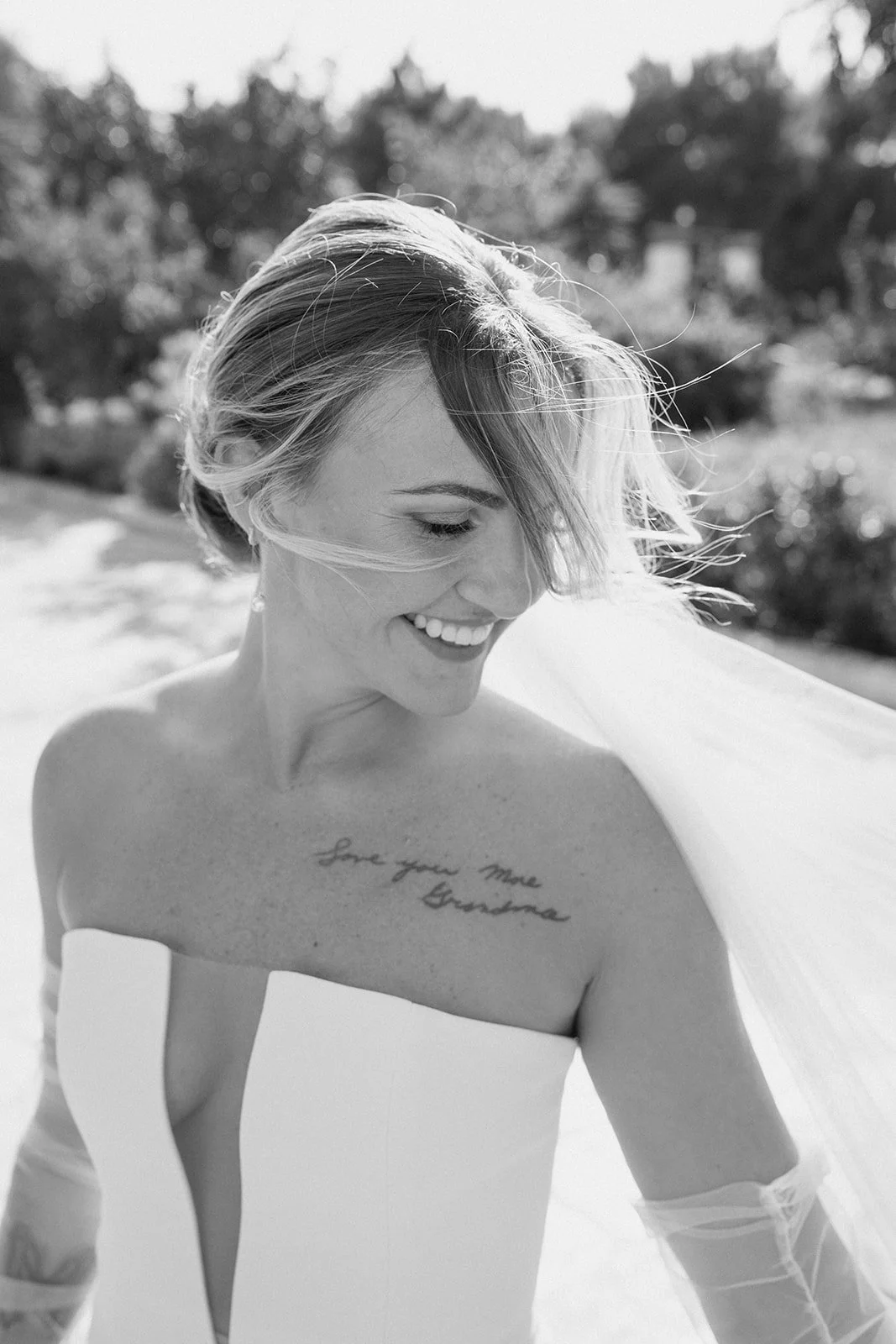 Black and white photo of a smiling woman with short hair, wearing a strapless dress, outdoors with trees in the background, and a tattoo on her chest that says 'Love you more Endless'.