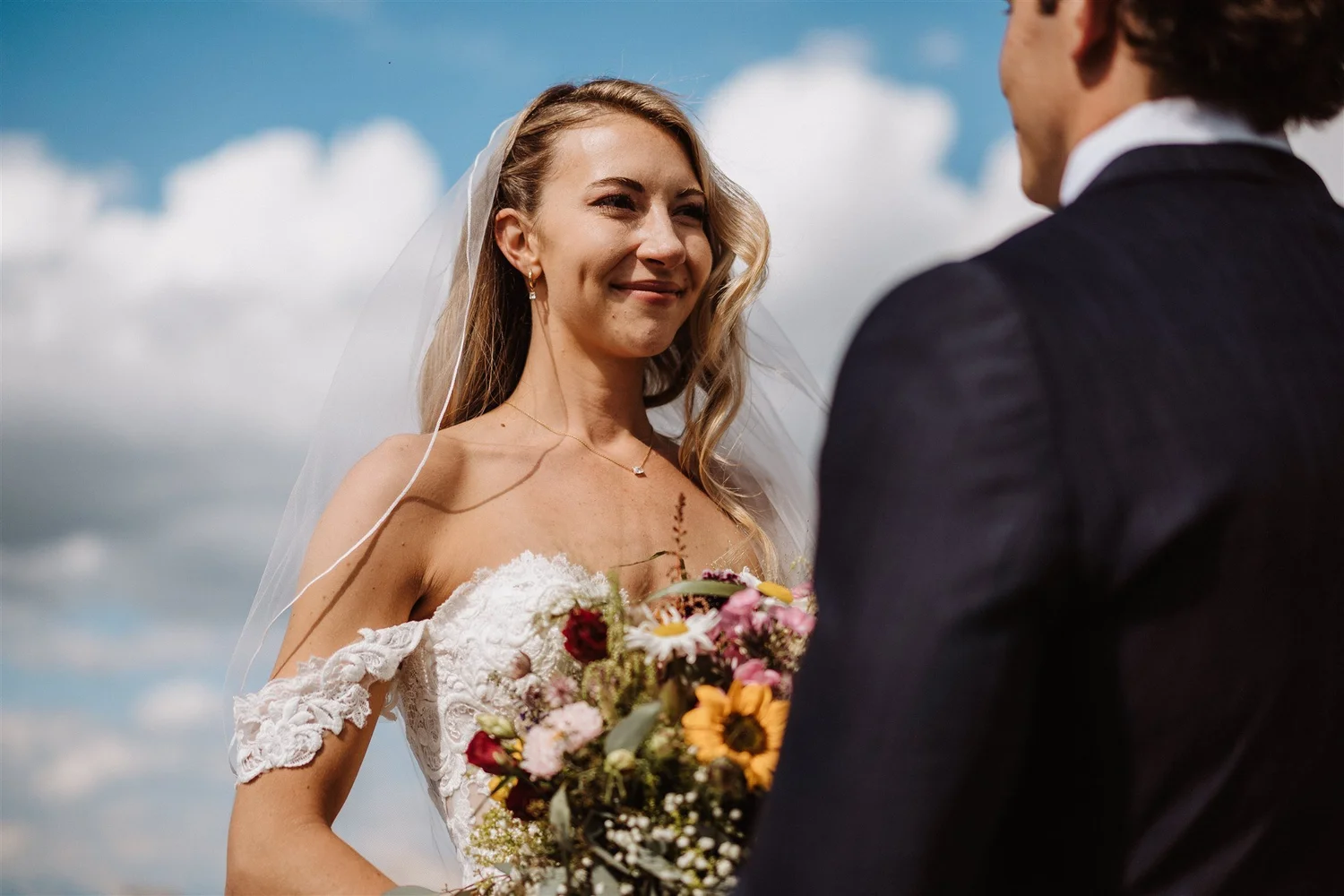 A bride and groom during her wedding vows outdoors under a cloudy sky. The bride is smiling and holding a bouquet of colorful flowers, while the groom, wearing a dark suit, faces her.