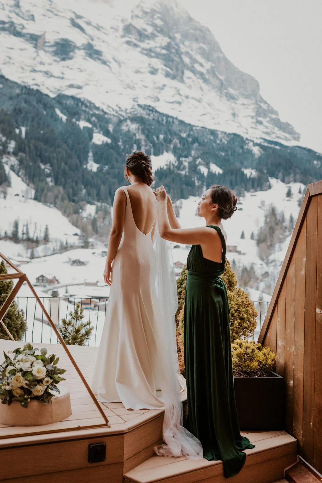 A bride in a white wedding dress standing on a wooden platform, while a woman in a dark green gown adjusts her dress, with snow-covered mountains and trees in the background.