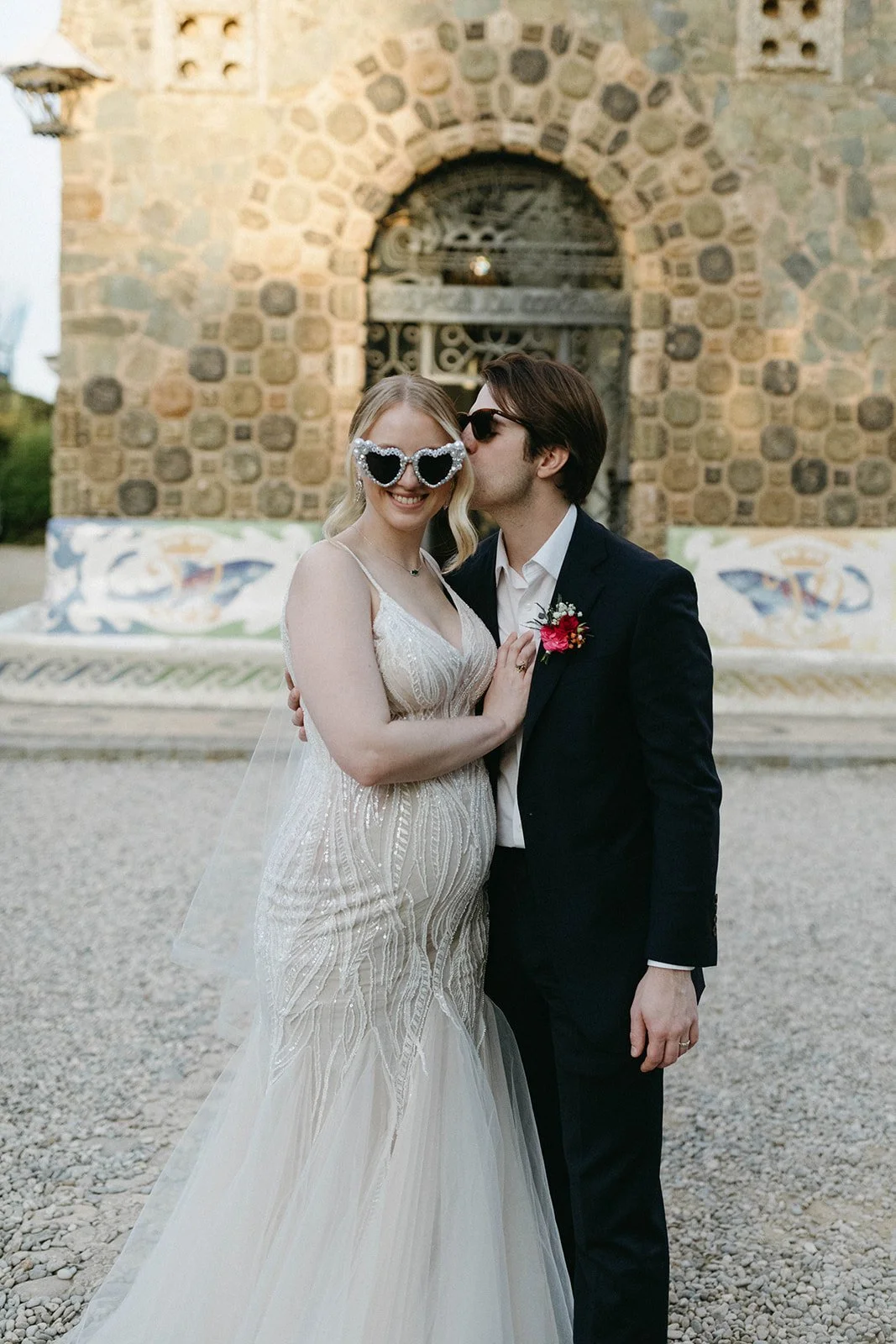 A bride and groom celebrating their wedding outdoors, with the groom kissing the bride on the cheek. The bride is wearing heart-shaped sunglasses and a white wedding dress, while the groom is in a dark suit with a boutonniere. They are standing in front of a stone wall with colorful murals and an arched window.