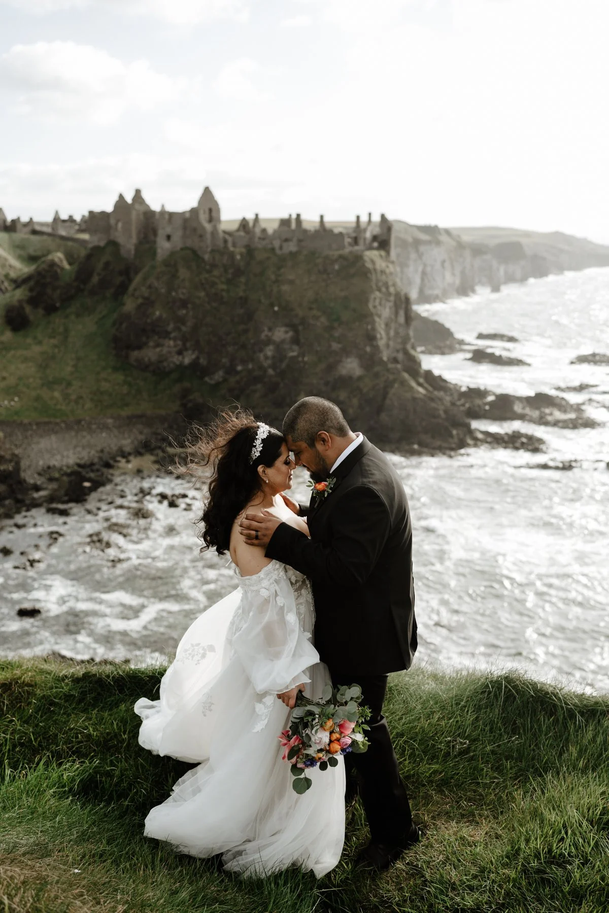 A bride and groom embrace on a grassy area near the ocean, with a historic castle on cliffs in the background, during their wedding photography session.